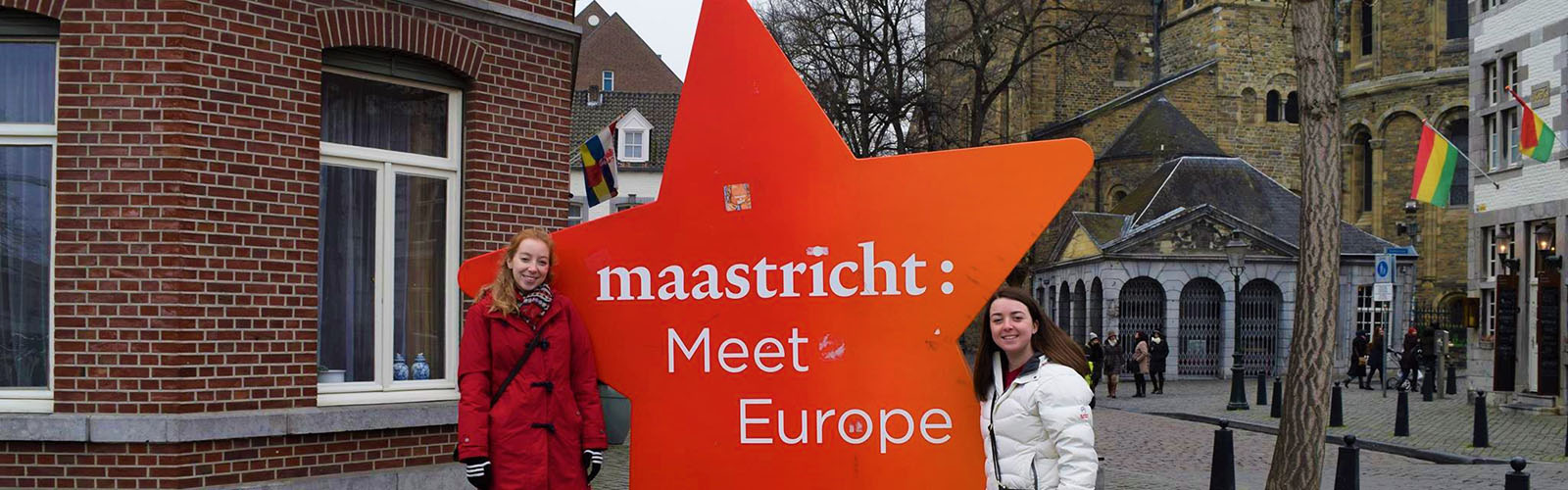 Two students stand in front of a red star-shaped sign that says "maastricht: Meet Europe". 